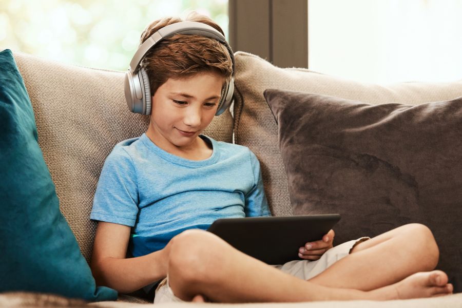 school aged boy sat on a sofa with an electronic tablet in his hand. He is wearing headphones.